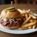 Delicious corned beef reuben sandwich with pickles and sauerkraut, elegantly plated with fries on a rustic wooden table.