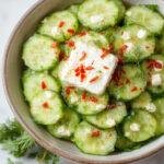 A vibrant, glossy cucumber salad arranged in a clear glass bowl. Smashed cucumber pieces are mixed with crumbles of white feta cheese and sprinkled with fresh dill. The salad has a bright green color with white feta accents, topped with a drizzle of oil and a few dill sprigs, presented on a rustic wooden table with natural lighting highlighting its freshness.