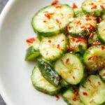 A vibrant bowl of Asian cucumber salad featuring thinly sliced crisp cucumbers coated in a tangy, spicy dressing with garnishes of chopped cilantro and sesame seeds, presented in a clean white bowl on a wooden table with a sprinkle of fresh herbs around.