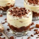 A close-up of a glass dessert cup filled with layered chocolate tiramisu, topped with cocoa powder and chocolate shavings. The layers are visible through the transparent glass, showcasing the creamy mascarpone and rich chocolate layers, with a dusting of cocoa on top for visual contrast.