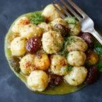 A vibrant plate of golden-brown gnocchi mixed with colorful sautéed vegetables such as cherry tomatoes, zucchini, and spinach, all coated in a glossy white wine lemon sauce, garnished with fresh herbs. The gnocchi have a slightly crispy exterior and soft interior, presented on a white ceramic plate with a lemon wedge and sprigs of basil for garnish.