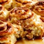 A baking dish filled with freshly baked peach cobbler cinnamon rolls topped with a dusting of powdered sugar, surrounded by fresh peach slices and cinnamon sticks.