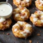 A close-up of golden-brown baked apple fritters with a glossy glaze, nestled on a rustic wooden plate. The fritters are round, slightly irregular in shape, with a crispy exterior and hints of cinnamon and apple visible inside. The glaze glistens under soft lighting, highlighting their tempting texture and warm color.
