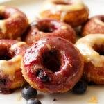A close-up of golden, glazed blueberry lemon curd donut holes garnished with fresh blueberries and lemon zest on a rustic plate.