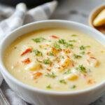 A warm bowl of creamy broccoli cheddar soup topped with shredded cheddar cheese and fresh chopped chives, served in a rustic white bowl with a golden spoon on a wooden table. The soup has a vibrant green and yellow appearance, with melted cheese and tender broccoli florets visible under a smooth, thick layer.