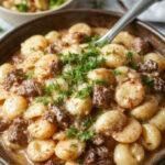 A close-up of creamy beef and shell pasta served in a rustic white bowl. The dish features tender beef chunks coated in a rich, creamy sauce dotted with al dente pasta shells. Garnished with finely chopped parsley for a splash of color, the sauce has a glossy texture highlighting the hearty and indulgent nature of the dish.