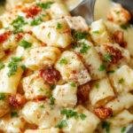 A bowl of creamy chicken pot pie pasta garnished with fresh herbs, featuring tender pasta mixed with chunks of chicken, vegetables, and a golden crust-like topping, served on a rustic wooden table.
