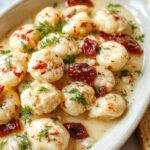 A close-up of a white ceramic plate featuring golden-brown gnocchi topped with a vibrant red sun-dried tomato sauce, garnished with fresh basil leaves, and drizzled with a creamy white sauce. The gnocchi appear soft and pillowy, with a crispy exterior, contrasted by the rich, colorful toppings and a rustic, inviting presentation.