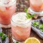 A tall, clear glass filled with vibrant pink whipped lemonade, topped with a swirl of whipped cream and a lemon wedge. The drink has a smooth, frothy texture with visible bubbles on the surface. The glass is garnished with a fresh mint sprig and colorful straws, set against a sunny summer backdrop with subtle bokeh effects.