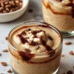 A tall glass of coffee mousse with a rich, velvety texture topped with whipped cream and a sprinkle of cocoa powder. The mousse is layered, showcasing its airy, light consistency with a deep coffee color. Garnished with a coffee bean and a drizzle of chocolate syrup, the presentation exudes elegance and indulgence, set against a sleek, neutral background.