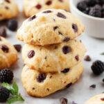 A close-up of golden-brown vegan blueberry lavender madeleines arranged elegantly on a white ceramic plate. The madeleines have a distinctive shell shape with a slightly domed top, speckled with fresh blueberries and tiny lavender buds. The surface is lightly textured with crisp edges and a soft, moist interior visible in some sliced pieces. The background features a subtle, rustic wooden table with a sprig of lavender and a few blueberries scattered nearby, enhancing the fresh and aromatic appeal.