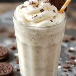 A tall glass filled with a rich, creamy Oreo milkshake topped with whipped cream and crushed Oreo cookies. The milkshake has a smooth, thick texture, with visible chunks of Oreos inside, and is garnished with a whole Oreo cookie on top. The background features a rustic wooden table and a colorful straw adds a playful touch.