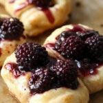 Golden-brown puff pastry twists with visible swirls of creamy white goat cheese and deep purple-black blackberry compote filling, arranged on a rustic wooden surface with a few fresh blackberries scattered around. The twists showcase flaky layers and a glossy finish, highlighting the contrast between the pastry and vibrant filling.