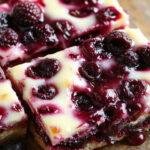 A plate of vibrant blueberry cheesecake bars topped with fresh blueberries and a sprinkle of powdered sugar, set on a summer-themed table.