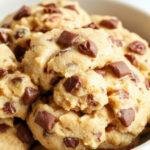 A close-up of a creamy, light tan edible cookie dough scooped into small mounds on a white plate, garnished with chocolate chips and a sprinkle of sea salt, with a rustic wooden backdrop and blurred baking utensils in the background.