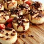 A colorful Epic Mini Cheesecake Board featuring various mini cheesecakes topped with fruits, sauces, and sprinkles, arranged artfully on a wooden platter.