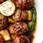 A plate of glazed Korean BBQ meatballs arranged neatly alongside roasted colorful vegetables including carrots, zucchini, and bell peppers. The meatballs have a glossy, caramelized surface with a rich brown color, garnished with sesame seeds and chopped green onions. The vegetables are roasted to perfection with slight charring, adding vibrant hues of orange, green, and purple, all presented on a rustic white plate on a wooden table.