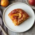 A vibrant peach upside-down cake featuring golden caramelized peach slices arranged elegantly on top, with a light, fluffy cake base visible around the edges. The cake is plated on a rustic wooden surface, with a few slices cut to reveal the juicy peach filling inside. The glossy caramel glaze enhances the warm, inviting appearance, and fresh peach slices are visible both on top and in the serving slices.