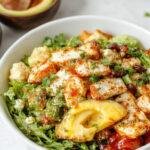 A vibrant bowl featuring sliced grilled chicken, fresh cherry tomatoes, crisp cucumber slices, red onion rings, and a creamy tzatziki sauce drizzled on top. The bowl is artfully arranged on a rustic wooden table, showcasing colorful ingredients and a sprinkle of fresh herbs for garnish.