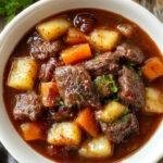 A rustic bowl of hearty red wine-braised beef stew filled with tender beef chunks, sliced potatoes, and carrots, garnished with fresh herbs.