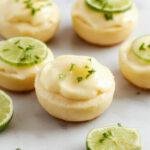 Miniature key lime pies with vibrant green lime filling topped with whipped cream and lime zest, presented on a rustic white plate with a golden crust edge, garnished with fresh lime slices and a sprig of mint. The photograph showcases the creamy texture of the lime filling contrasted with the crisp crust and the bright, inviting colors.