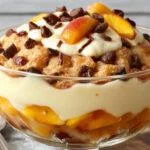 A layered peach cobbler trifle in a clear glass, showcasing ripe peach slices, golden cobbler topping, and smooth vanilla pudding, topped with whipped cream and a sprig of mint.