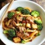A close-up of a bowl of homemade beef and broccoli lo mein showcasing tender slices of beef, vibrant green broccoli florets, and glossy noodles garnished with sesame seeds and chopped green onions, presented in a rustic ceramic bowl on a wooden table.