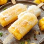 Colorful homemade coconut mango popsicles with vibrant orange and white hues, garnished with fresh mango slices and coconut flakes, set in a summer-themed background.