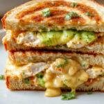 A close-up of a crispy chicken avocado melt sandwich showing melted cheese, tender chicken slices, ripe avocado, and toasted bread garnished with fresh herbs.