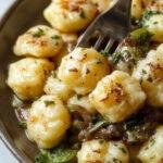A close-up of golden-brown gnocchi topped with melted cheese and caramelized onions, garnished with fresh herbs, arranged on a rustic plate with a rich, creamy sauce visible around the edges.