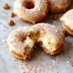Golden-brown Italian doughnuts filled with creamy custard, dusted lightly with powdered sugar, arranged on a white plate with a rustic background, showcasing their soft, airy texture and luscious filling.