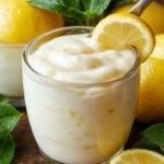 A jar of vibrant yellow Italian Lemon Spread surrounded by fresh lemon slices and sprigs of mint, on a rustic wooden table.