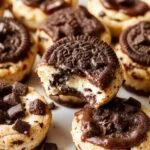 Close-up of golden-brown Oreo cheesecake cookie cups filled with smooth, creamy cheesecake filling topped with crushed Oreo pieces. The cookie cups are neatly arranged on a rustic wooden surface, with a few Oreos scattered around, highlighting their layered texture and inviting appearance.