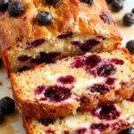 A fresh Lemon Blueberry Yogurt Loaf sliced on a cake stand, revealing its moist interior dotted with blueberries and a bright lemon glaze on top.