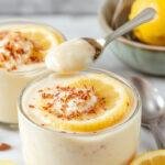 A close-up of a glass jar filled with lemon-colored chia pudding topped with a swirl of whipped cream and garnished with fresh lemon slices and mint leaves. The pudding has a creamy, textured appearance with visible chia seeds, presented on a rustic wooden surface alongside a lemon wedge and a small spoon.