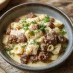 A steaming bowl of creamy ground beef and pasta topped with chopped herbs, showcasing tender pasta coated in a rich, white sauce and bits of browned ground beef, garnished with fresh parsley.