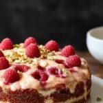 A layered Pistachio Raspberry Cake with vibrant pink raspberry filling, green pistachio frosting, and fresh raspberries on top, elegantly presented on a white plate.