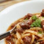 A bowl of creamy beef noodles featuring tender slices of beef, swirled in a rich, white sauce, topped with fresh herbs and served with a side of vibrant vegetables. The noodles are glossy and coated evenly, garnished with a sprinkle of green herbs, presented on a rustic wooden table.