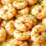 A close-up of creamy garlic shrimp arranged on a white plate. The shrimp are plump, coated in a luscious, buttery garlic sauce with visible minced garlic and herbs. Garnished with fresh parsley, the dish is complemented by a side of lemon wedges and a sprinkle of black pepper, creating a vibrant and appetizing presentation with a glossy texture.