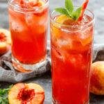 A tall glass of Sweet Summertime Peach Lemonade garnished with fresh peach slices and lemon wedges, surrounded by whole peaches and lemons on a sunny outdoor table.
