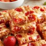 A close-up of golden-brown strawberry lemon blondies topped with fresh strawberry slices and a drizzle of lemon glaze, arranged on a rustic white plate. The blondies reveal a soft, chewy interior with bits of strawberries visible throughout, accented by vibrant red and yellow fruit toppings that add a fresh, colorful contrast.