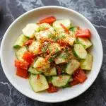 A colorful Thai cucumber salad served in a clear glass bowl, featuring thinly sliced bright green cucumbers coated with a tangy reddish-orange chili and lime dressing, garnished with fresh cilantro and dotted with red chili slices. The salad has a fresh, crisp texture with a glossy sheen from the dressing, contrasting the vibrant green cucumbers with the fiery red and orange accents.