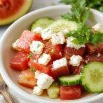A vibrant salad featuring juicy red watermelon cubes, crisp green cucumber slices, and crumbled white feta cheese, all arranged on a white plate with fresh mint leaves. The colorful ingredients are complemented by a light drizzle of vinaigrette, creating a fresh and inviting presentation with a summery theme.