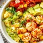 A vibrant bowl filled with plump, pink shrimp arranged atop sliced creamy avocado and bright mango salsa, garnished with fresh cilantro and lime wedges, all presented on a white plate against a colorful tropical backdrop.