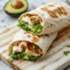 A fresh, colorful chicken avocado wrap on a white ceramic plate, cut diagonally to reveal vibrant layers of grilled chicken, creamy avocado, crisp lettuce, and red bell pepper, wrapped in a soft whole wheat tortilla with slight grill marks, garnished with lime zest and microgreens, natural daylight, shallow depth of field, minimal styling, lower third of image empty.