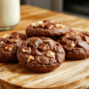 A batch of thick, chewy chocolate walnut cookies with golden-brown edges, cracked surfaces revealing melted chocolate chips and walnut pieces, dusted lightly with powdered sugar, sitting on a rustic wooden board beside a glass of milk