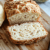 A golden-brown, dome-shaped loaf of keto yogurt bread on a rustic wooden cutting board, sliced to reveal a soft, airy crumb with tiny air pockets and a slight crust. A drizzle of melted butter glistens on the top slice, with fresh rosemary sprigs and coarse sea salt scattered nearby on the board.