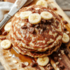 Stack of fluffy banana oatmeal pancakes on a white plate with golden edges, banana slices and berries on the side, oats visible in the batter, light maple drizzle, warm natural lighting, rustic wooden table, inviting and wholesome.