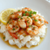 Plate of golden buttery garlic shrimp arranged atop fluffy white rice, vibrant green parsley, lemon wedges, and a glossy garlic butter sauce, with cracked pepper on a light wooden table.