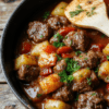 A rustic skillet filled with browned beef chunks, golden potato cubes, onion and red bell pepper in a glossy tomato broth, garnished with chopped parsley. The pan rests on a wooden table with a light olive oil drizzle and a pinch of paprika, exuding cozy home-cooked comfort.